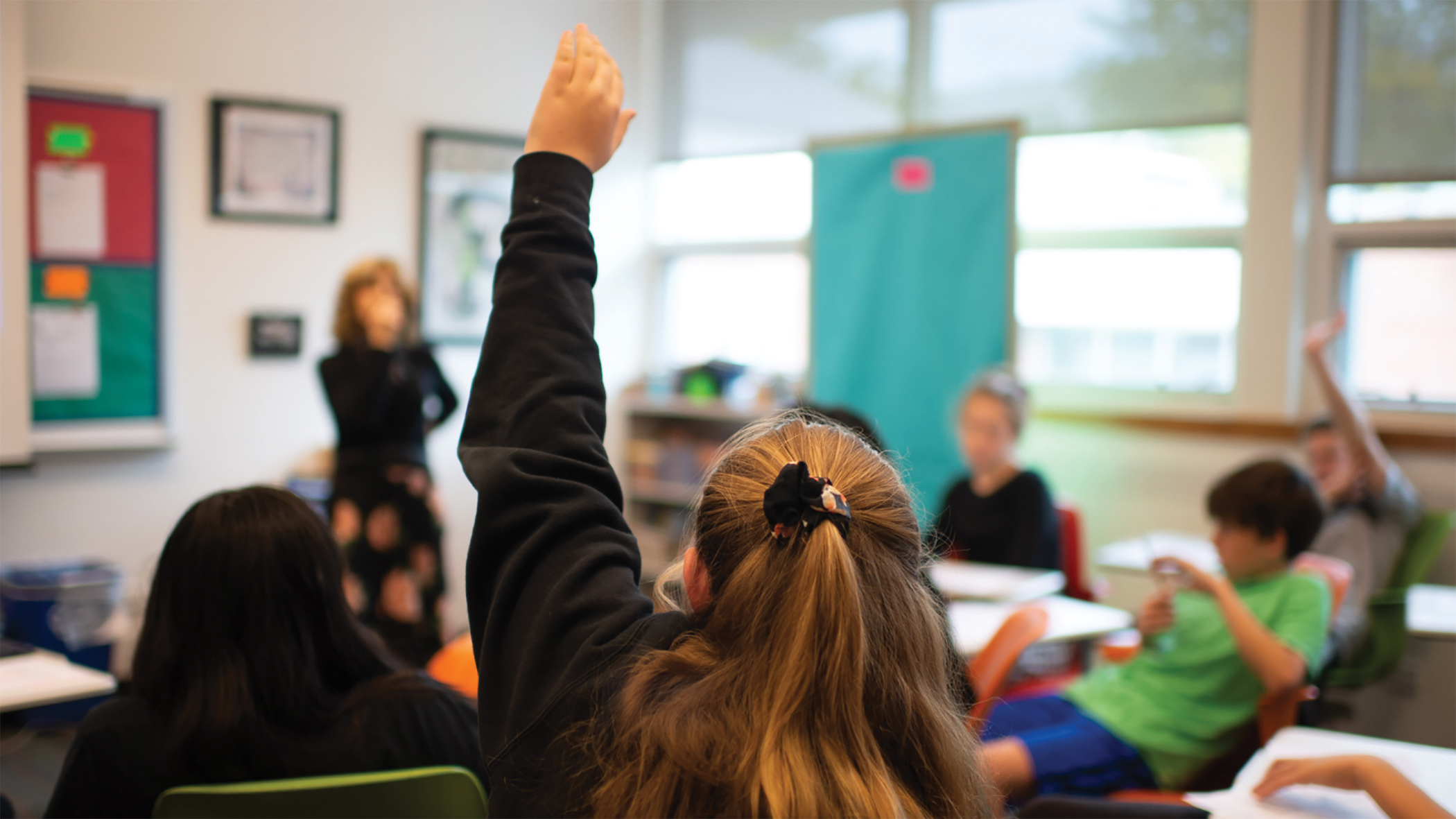 Closeup of student raising hand in classroom. Other students and teacher at front of classroom.