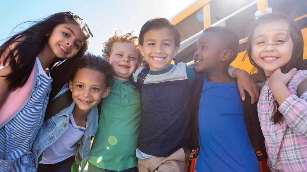 A group of diverse children with arms around each other standing in front of a school bus smiling