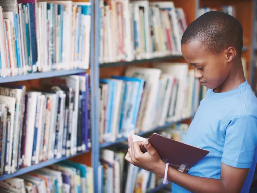 A young brown boy standing in a library reading a book