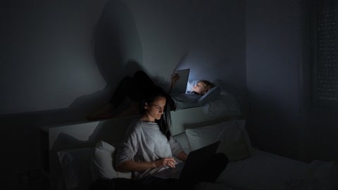 Mom in bedroom on laptop with child in bed on phone